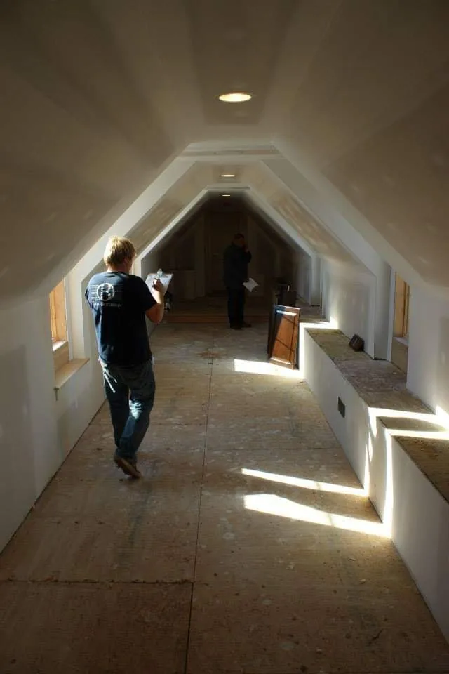 The same attic hallway before the woodwork, with bare drywall and two people measuring.